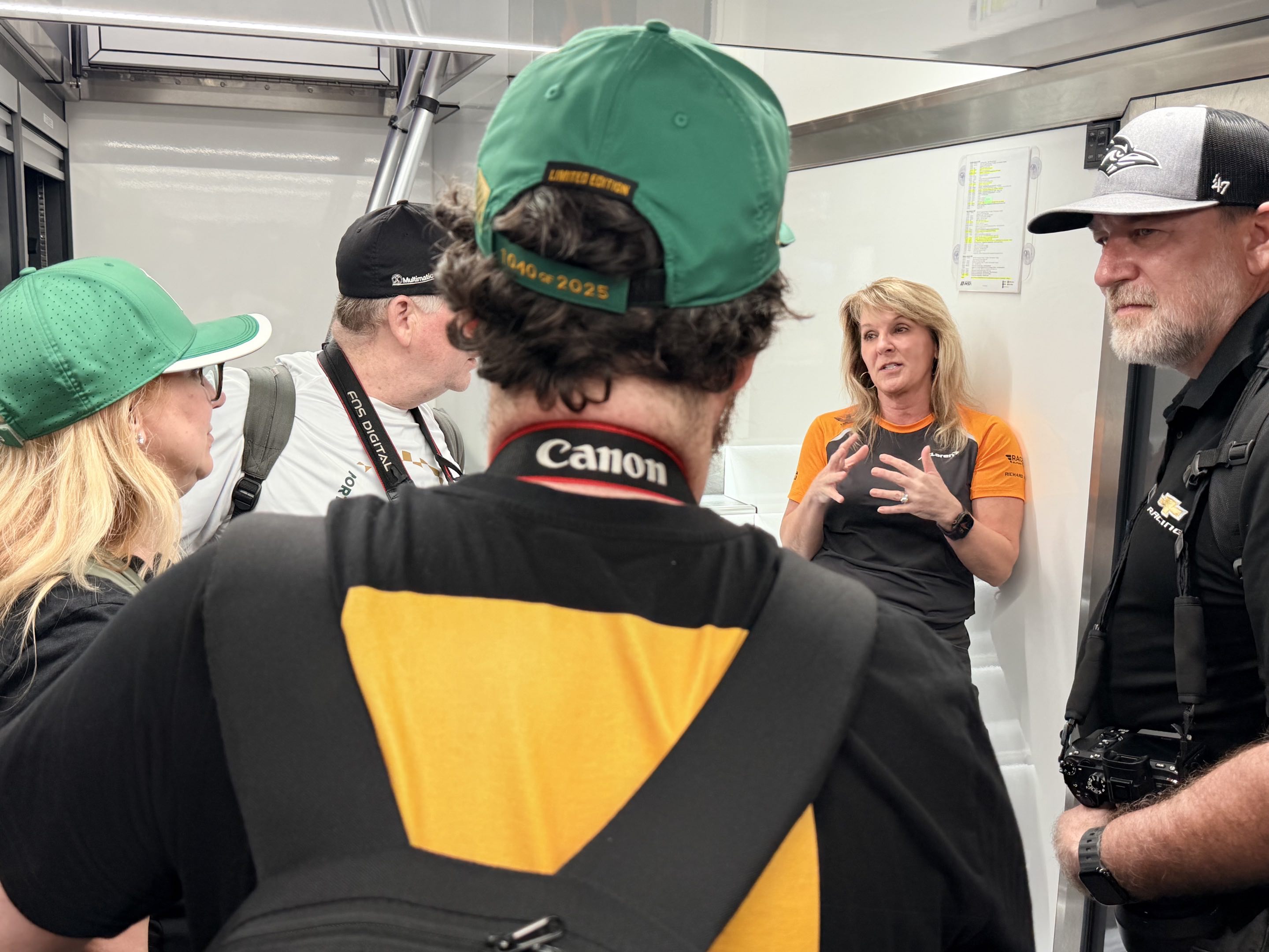 A group of people standing in a circle in the close confines of a hauler listening to a woman in a McLaren Racing shirt explain things